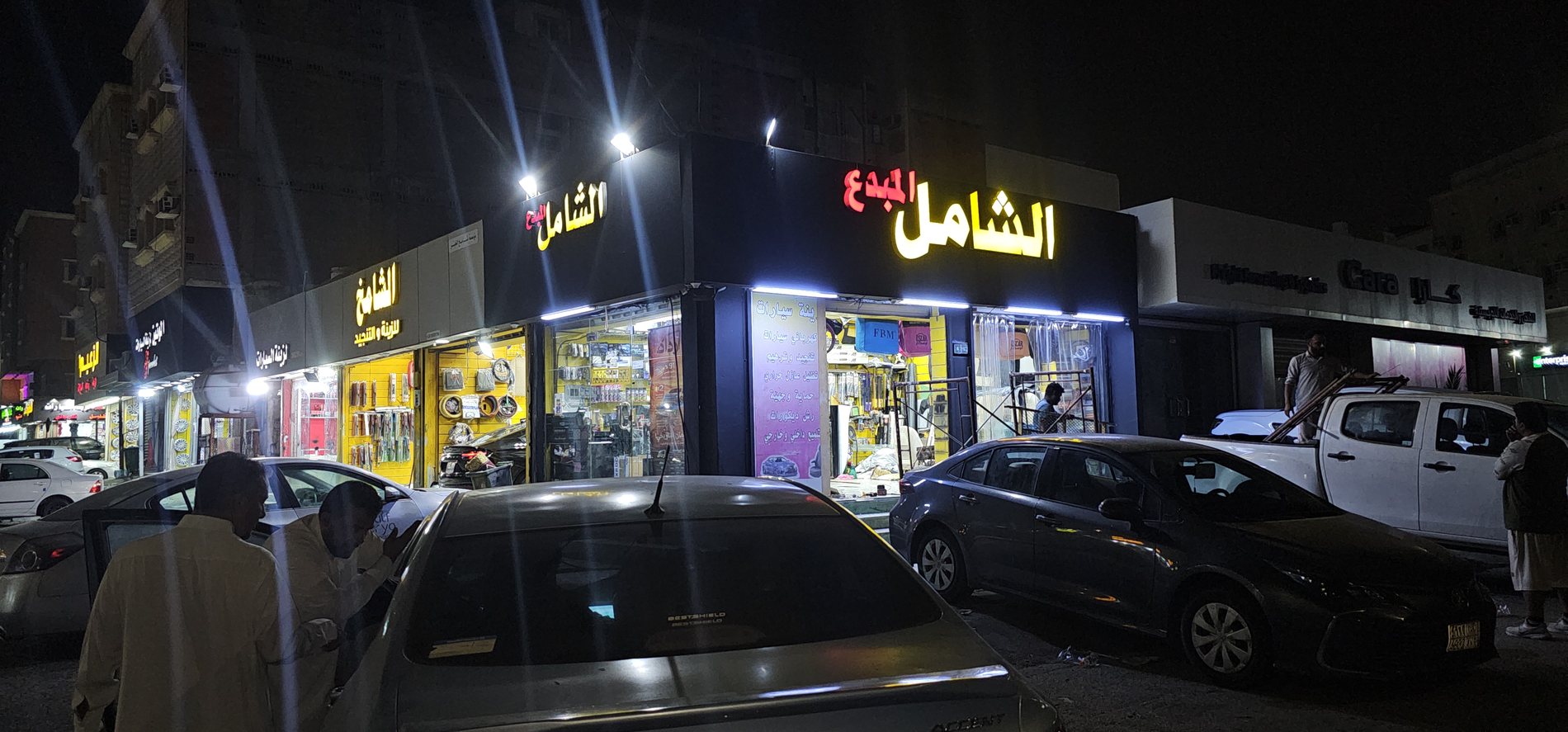 واجهة محل الشامل لزينة السيارات ليلًا، مع لوحة المحل المضيئة